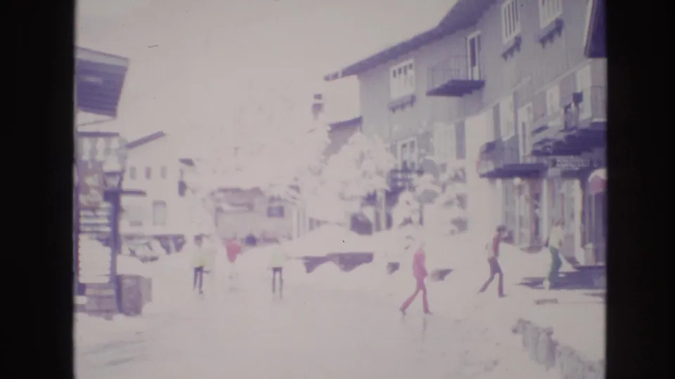 1974 Vail, Colorado home-movie frame showing people in winter clothing crossing and walking along a snowy resort street beside wooden buildings and balconies.