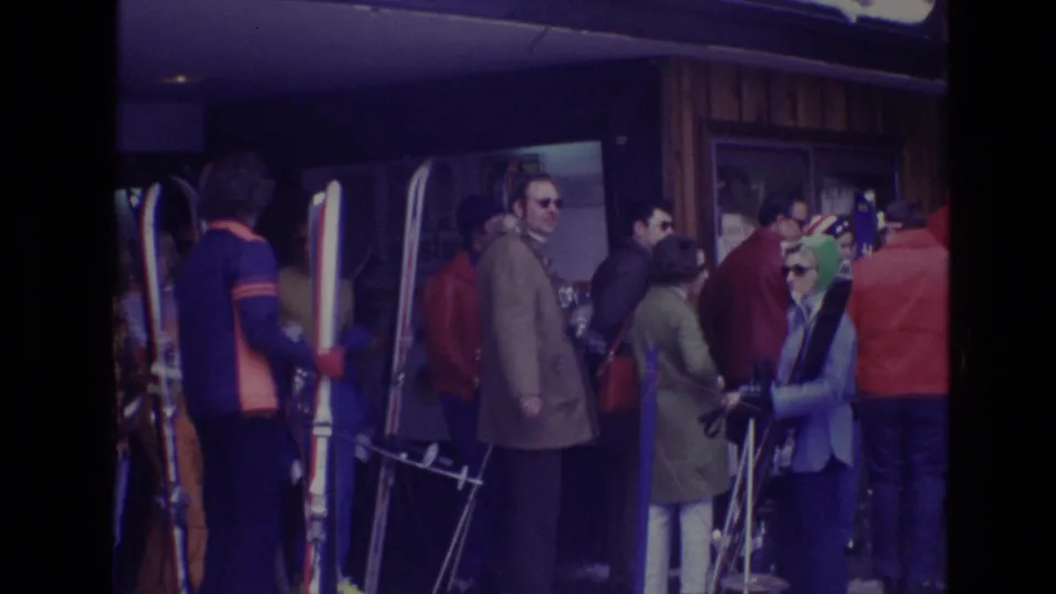 Visitors at a Vail ski resort in 1974 wait close together with long skis, poles, sunglasses, and colorful winter coats outside a wood-sided building.