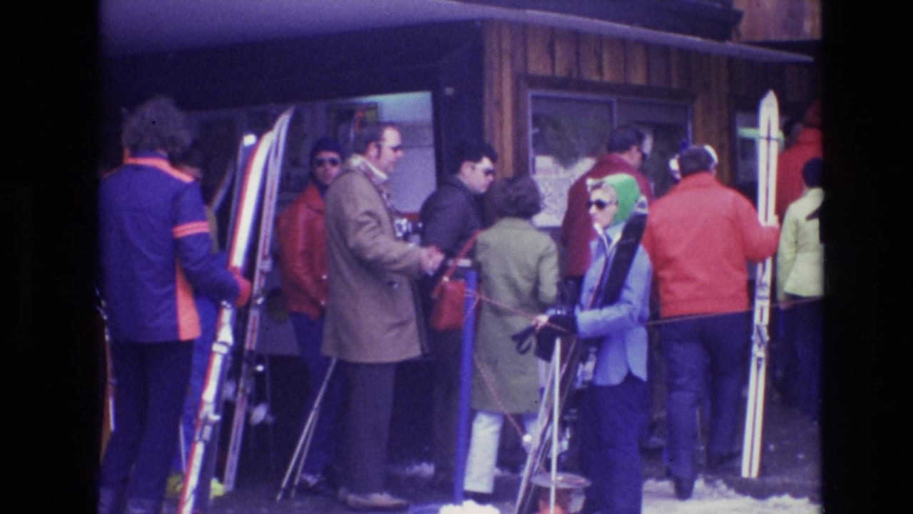 Adults in 1970s winter clothing wait near a Vail lodge entrance, with skis, poles, sunglasses, and snow visible around the queue.