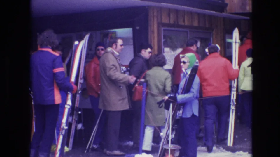 Adults in 1970s winter clothing wait near a Vail lodge entrance, with skis, poles, sunglasses, and snow visible around the queue.