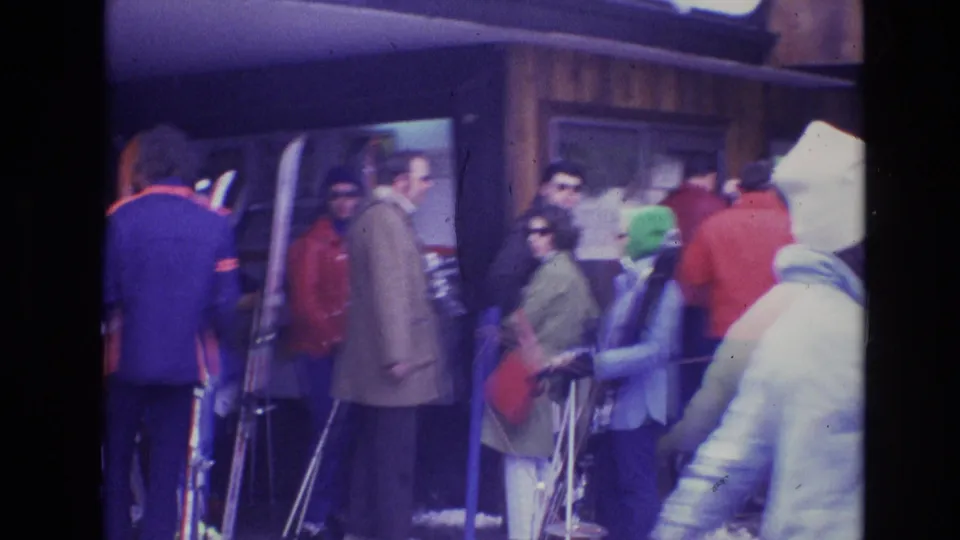A handheld 8mm frame from the 1974 Vail ski scene shows skiers waiting with poles near a wooden lodge as a bundled figure crosses the foreground.