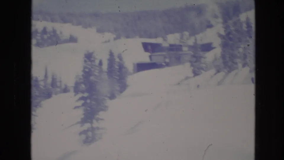 1974 Vail, Colorado 8mm frame showing a snow-covered slope, evergreen trees, and distant resort or lift-area buildings viewed from a visitor perspective.