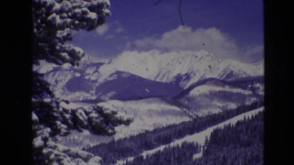 1974 Vail, Colorado 8mm frame showing snow-covered mountain ridges, forested slopes, and a dark evergreen branch in the foreground beneath a blue winter sky.