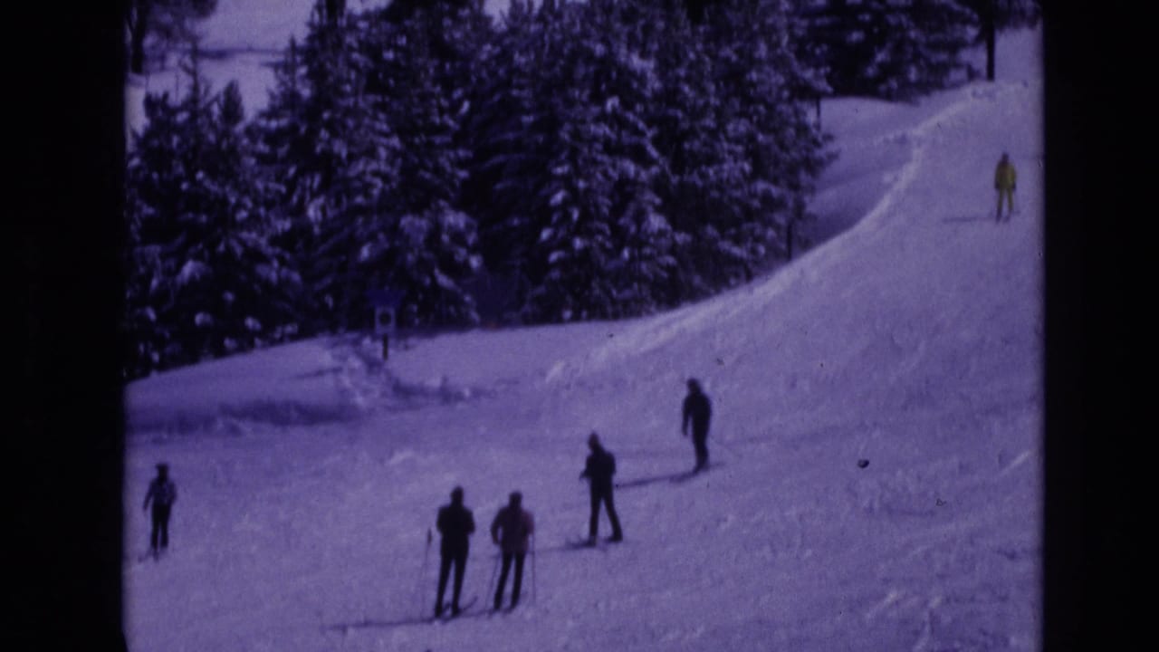 1974 Vail, Colorado frame with several skiers spread across a snowy resort slope bordered by dense evergreen trees.