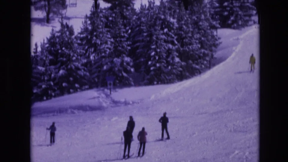 1974 Vail, Colorado home-movie frame showing skiers in dark and bright winter coats standing and skiing near a snowy evergreen backdrop.