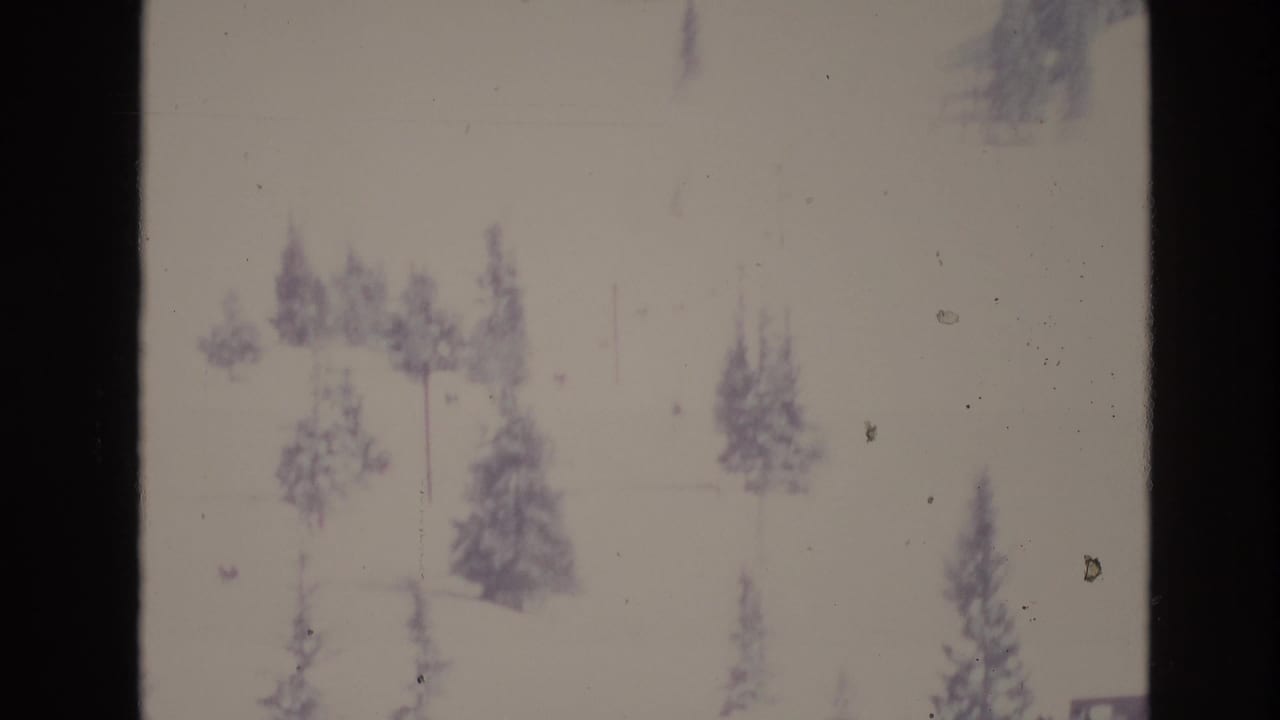Soft, overexposed 8mm frame of a snowy Vail-area slope in 1974 with scattered evergreen trees and faint ski-lift structures visible through the bright snow.