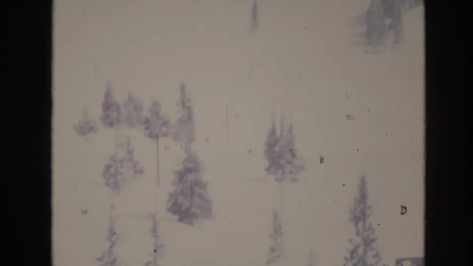 Soft, overexposed 8mm frame of a snowy Vail-area slope in 1974 with scattered evergreen trees and faint ski-lift structures visible through the bright snow.