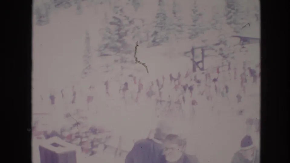Wide 8mm view of a busy 1974 Vail snowfield with groups of visitors moving near trees and resort structures, with a foreground figure partly visible at the lower edge.