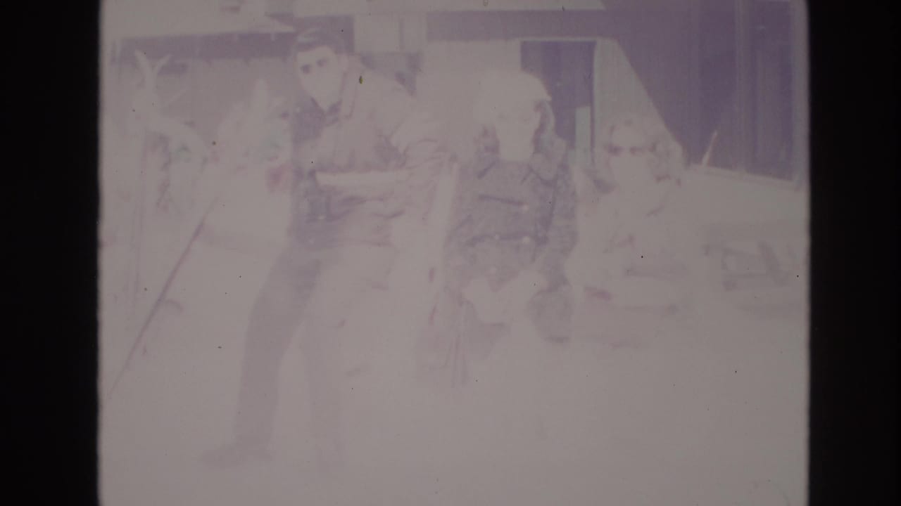 1974 Vail home-movie frame showing adults in winter coats standing in overexposed snow near a lodge or resort building.