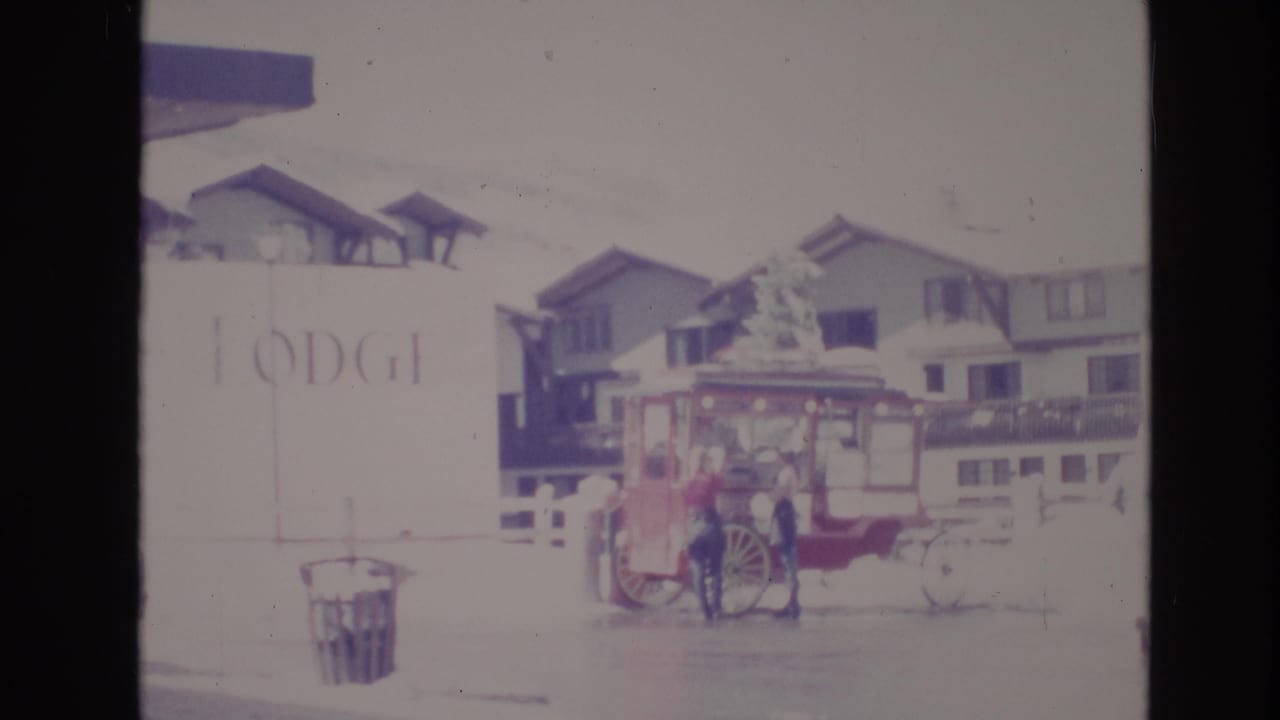 1974 Vail, Colorado 8mm frame showing a red canopy cart with large spoked wheels outside snow-covered alpine-style lodge buildings, with a person standing nearby.