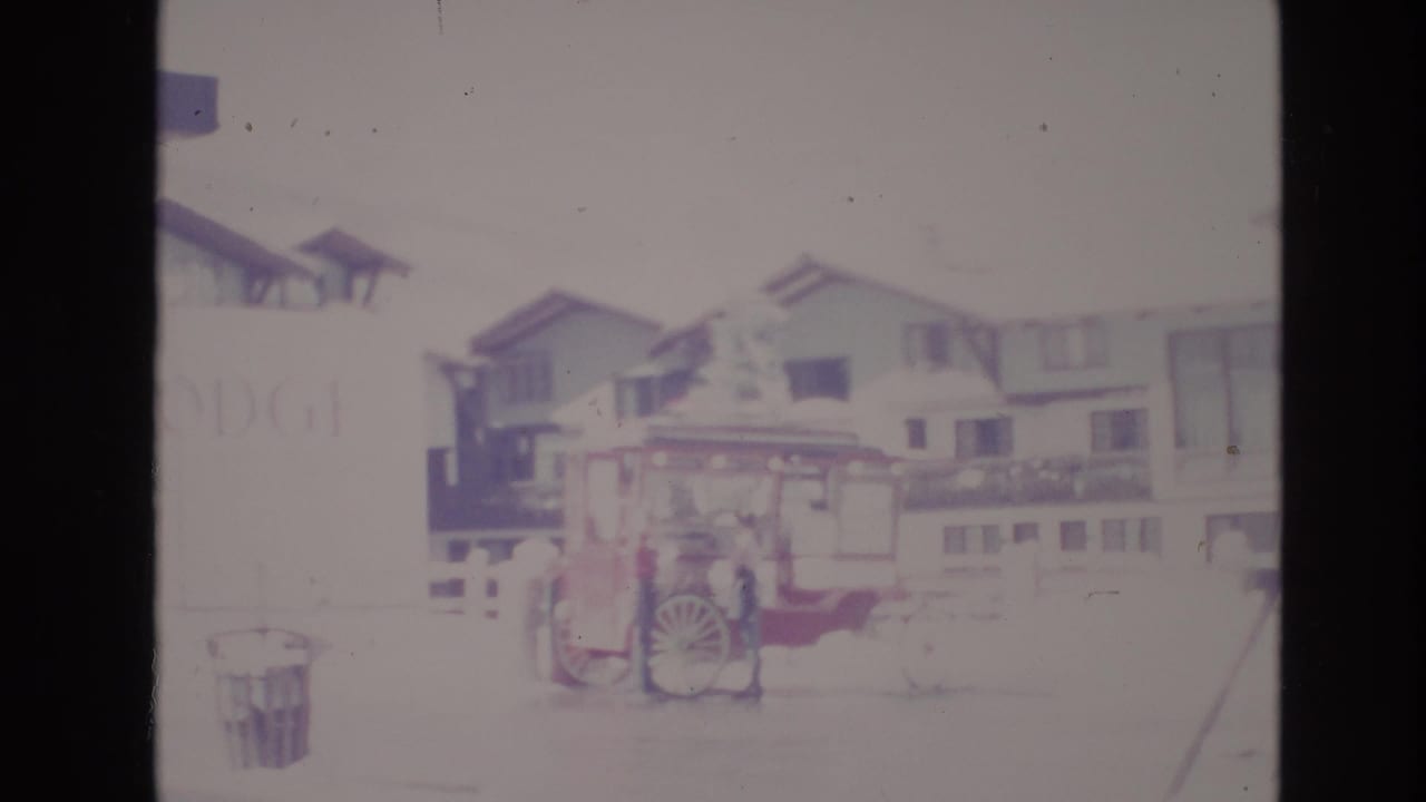 Soft, overexposed 1974 Vail, Colorado frame showing the red cart parked in front of snowy resort buildings with a faint sign at left.