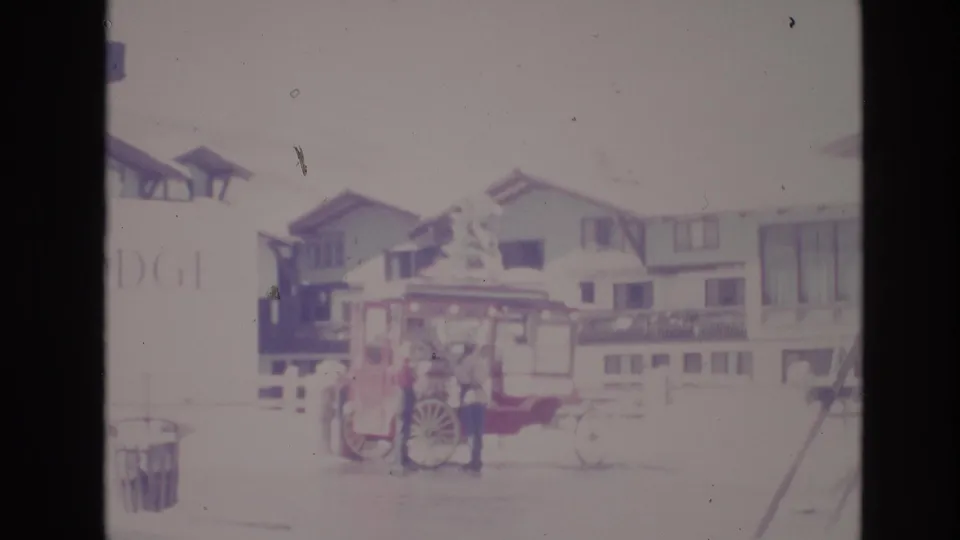 1974 Vail, Colorado home-movie frame with a person standing beside a red horse-drawn or vendor-style cart, snowy ground, and alpine lodge buildings in the background.