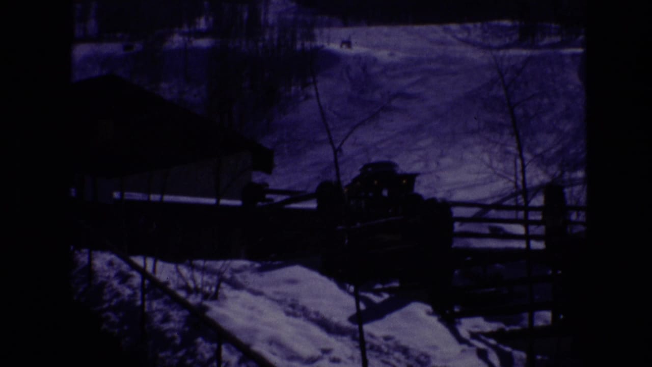 1974 Vail, Colorado home-movie frame showing a snow-covered resort area, a low modern building, wooden fencing, and a parked vehicle under heavy blue-purple tint.