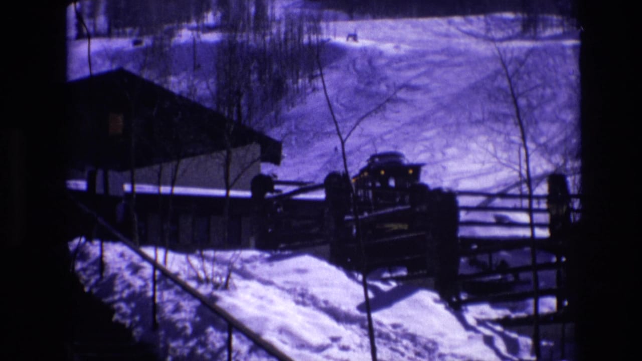 1974 Vail, Colorado frame with a parked vehicle beside wooden fencing, a snowbank, bare trees, and the edge of a lodge-like resort building.