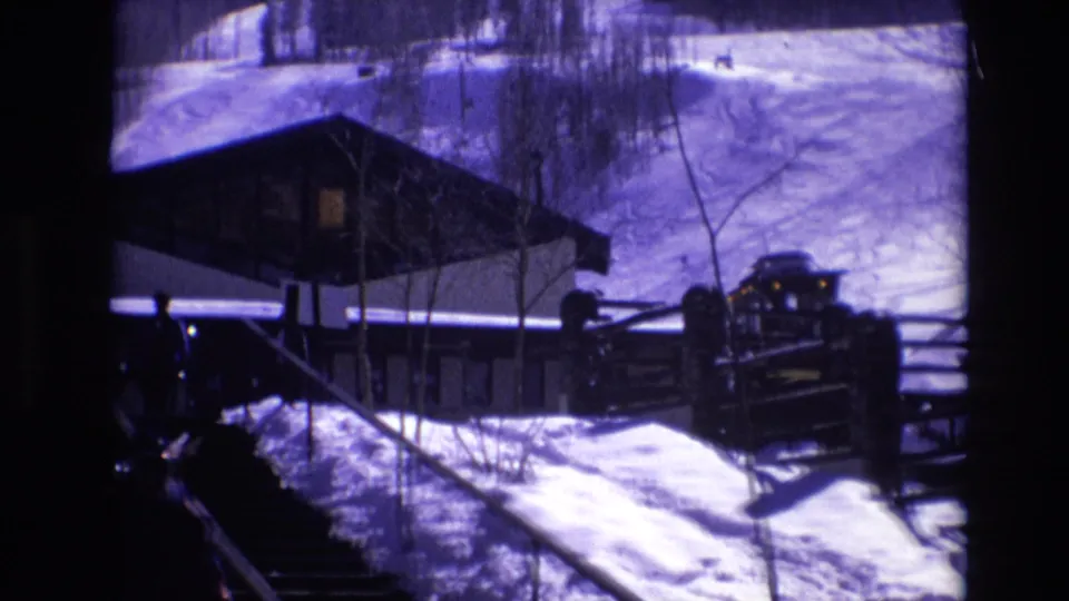 1974 Vail, Colorado 8mm frame showing exterior stairs in deep snow, a modern resort building, a parked vehicle, fencing, and bare winter trees.