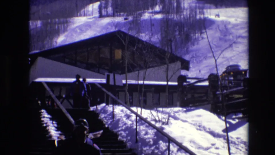 1974 Vail, Colorado home-movie frame showing a person in winter clothing on snow-covered stairs near a lodge-like building, wooden fencing, and a parked vehicle.