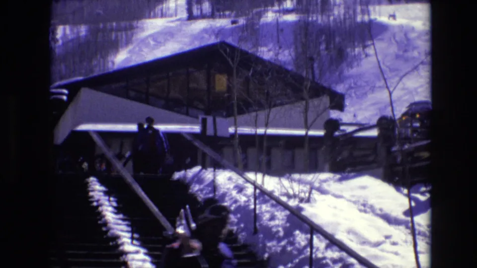 1974 Vail, Colorado 8mm frame showing a snow-covered stairway and railings leading toward a modernist hilltop resort building with winter visitors nearby.