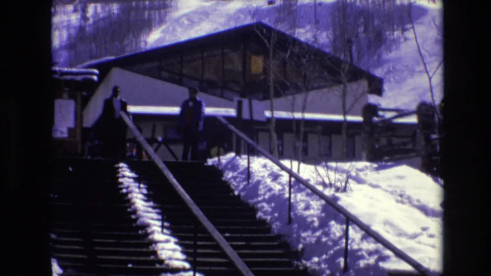 1974 Vail, Colorado 8mm frame showing winter visitors on a broad outdoor stairway with snow banks and a modernist hilltop building behind them.