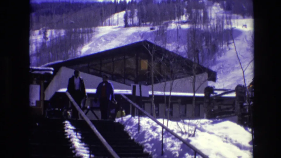 1974 Vail, Colorado frame showing two distant visitors on snow-lined stairs leading down from a modernist building on an alpine bluff.
