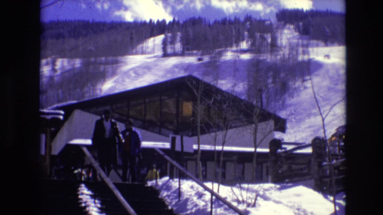 Two people in winter clothing stand on steps beside a modernist ski lodge in Vail, Colorado, with snow-covered slopes and evergreen forest visible in the background.