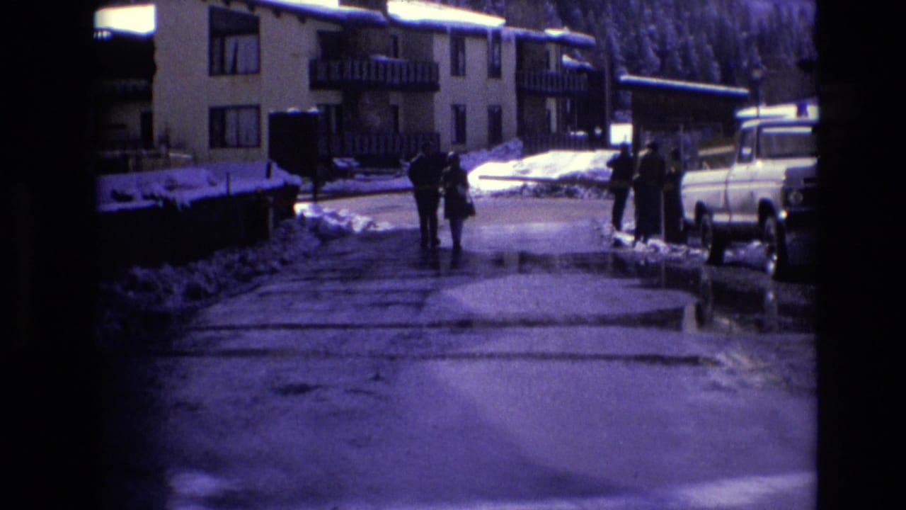 People in winter clothing move along a wet road in Vail, Colorado, near multi-story resort buildings, snowbanks, and a light-colored 1970s pickup truck.