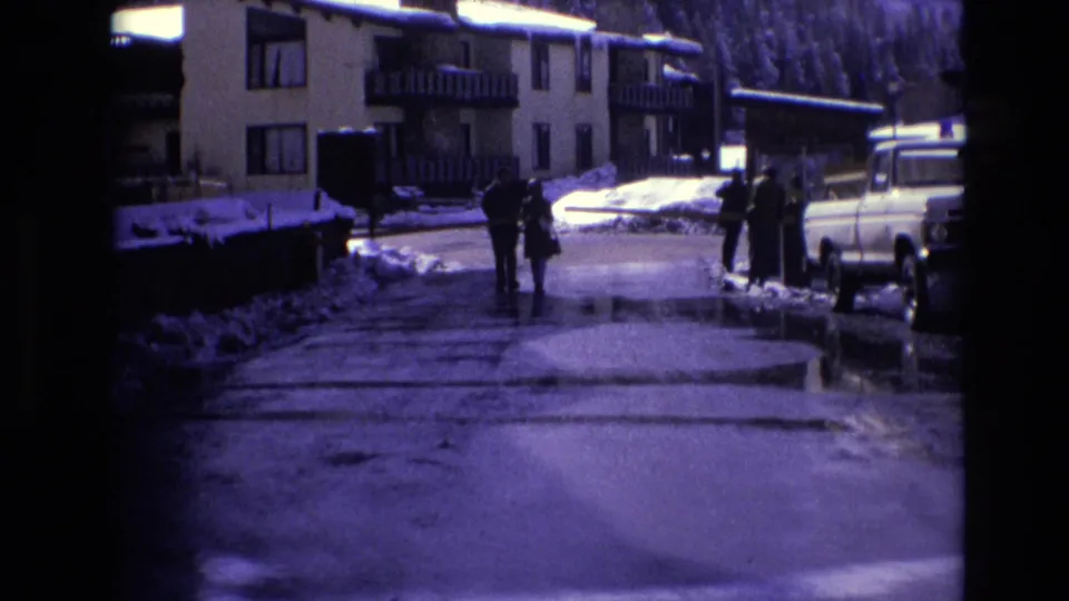 People in winter clothing move along a wet road in Vail, Colorado, near multi-story resort buildings, snowbanks, and a light-colored 1970s pickup truck.
