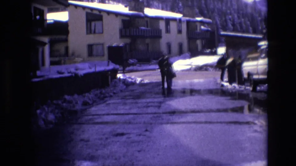 A handheld 8mm view shows a wet snow-lined road in Vail, Colorado, with bundled visitors near resort buildings and parked vehicles during a winter holiday scene.