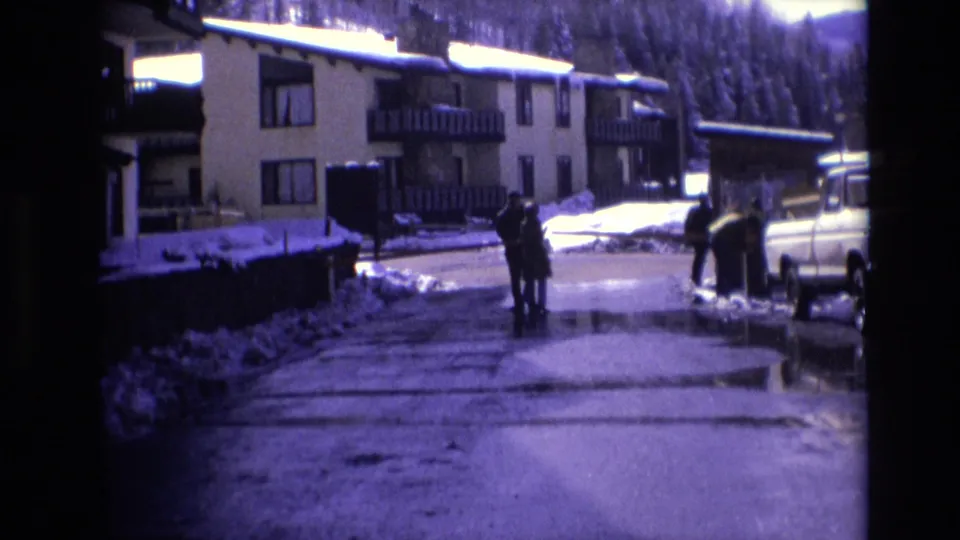 People stand and walk on a wet resort road in Vail, Colorado, framed by snowbanks, chalet-like lodging, wooded slopes, and a light-colored pickup truck.