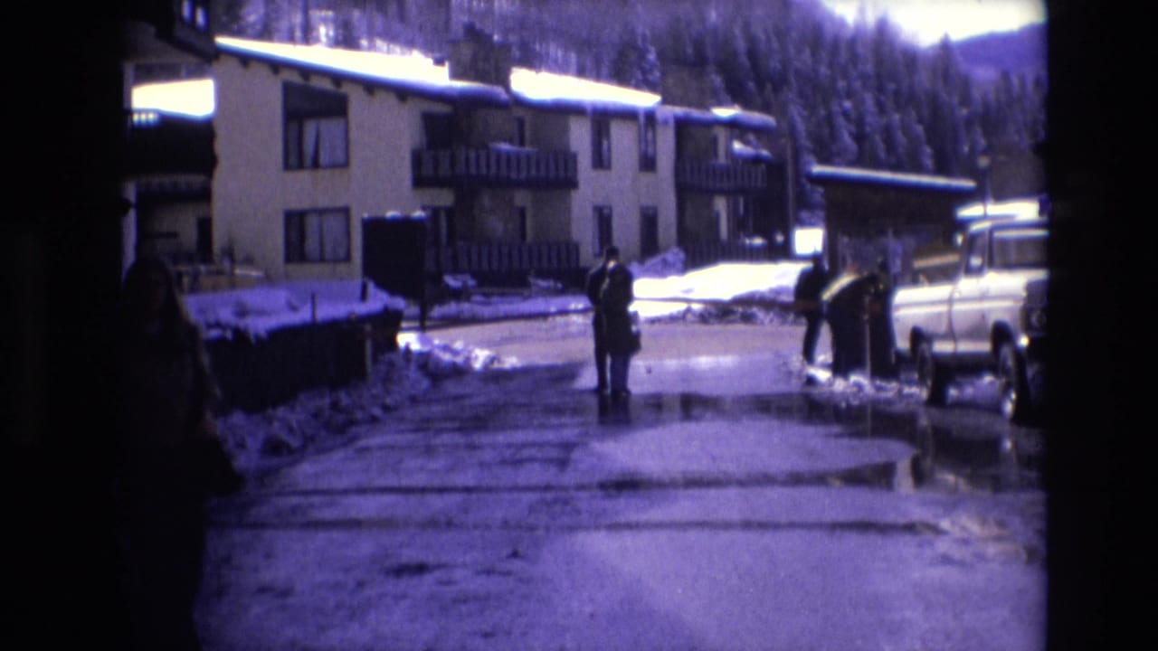 Two bundled people on a snowy resort road near apartment buildings and a white pickup truck.