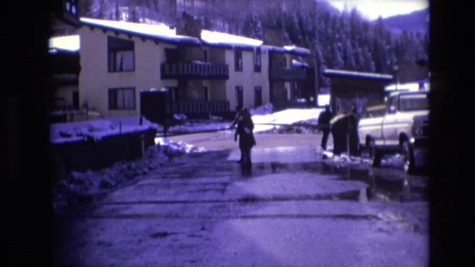 In 1974 Vail, bundled pedestrians move along a wet snowy road beside apartment-style lodging, a parked white pickup truck, and dark evergreen trees beyond the buildings.