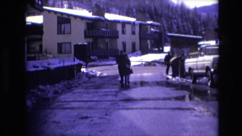 Two people in winter coats walk together through a snowy Vail resort road in 1974, with apartment balconies, snowbanks, a white pickup truck, and forested hills in the background.