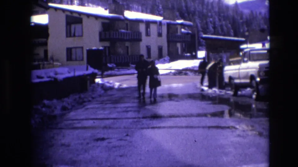 Two bundled people walk side by side on a slushy road in 1974 Vail, with snow-covered apartment buildings, a parked pickup truck, and evergreen hills behind them.