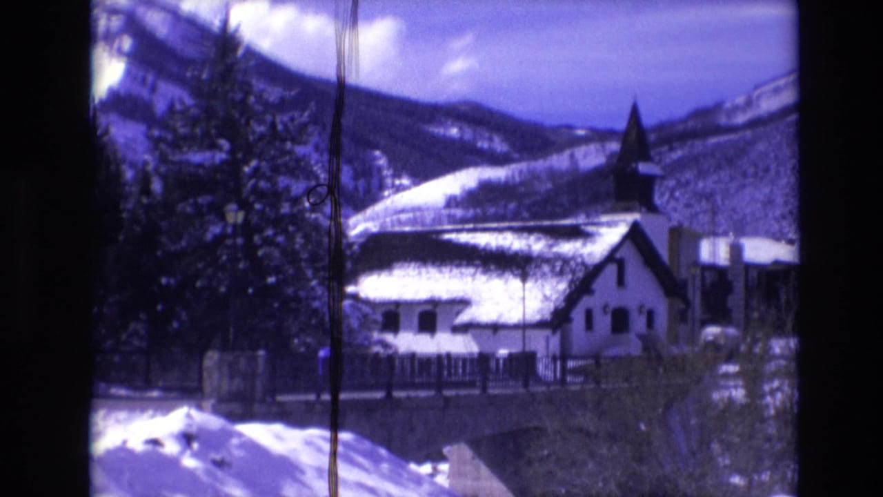 1974 Vail, Colorado home-movie frame showing a snow-covered church with a dark steeple, evergreen trees, fencing, and mountain slopes in the background.