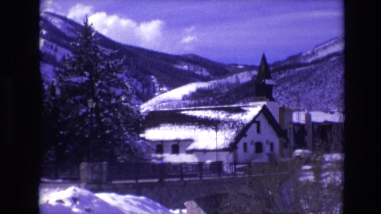 Church with snow on the roof beside evergreens and mountain ridges.