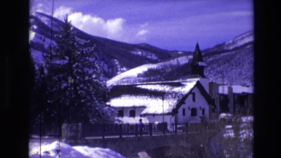 1974 Vail winter frame showing the church and steeple partly framed by an evergreen, with snowbanks, mountains, and visible 8mm film wear.
