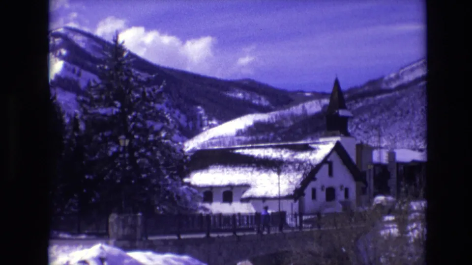 A 1974 Vail, Colorado 8mm frame of a snow-covered church and steeple near a low bridge or wall, with trees and mountain ridges behind it.