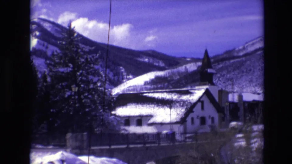 1974 Vail, Colorado 8mm frame showing a snow-covered church with a dark steeple, evergreen trees, and mountain slopes in the background.