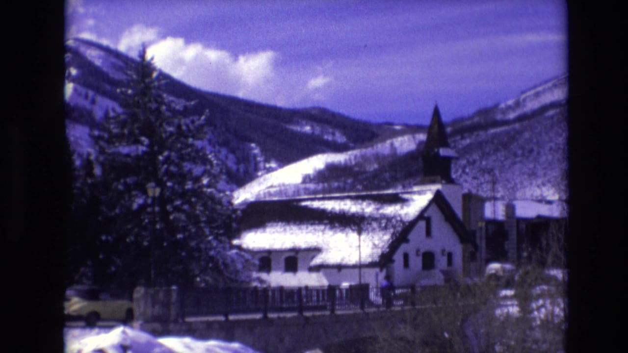 1974 Vail, Colorado 8mm frame with the snow-covered church, mountain backdrop, evergreen trees, and a yellow car moving along the road in front.