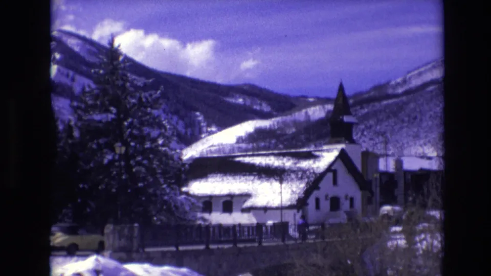 1974 Vail, Colorado 8mm frame with the snow-covered church, mountain backdrop, evergreen trees, and a yellow car moving along the road in front.