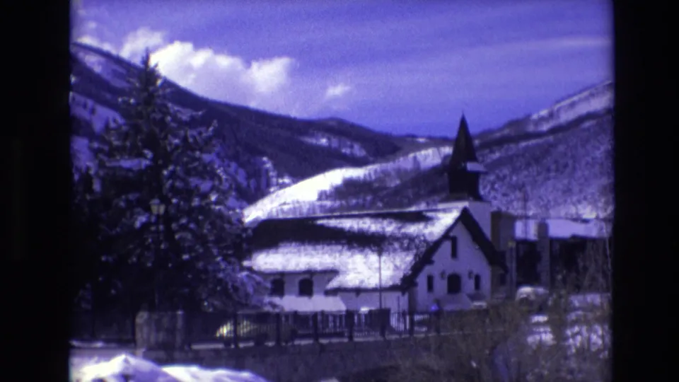 1974 Vail, Colorado 8mm frame holding on a white church with dark steeple, snow-covered rooflines, evergreens, and a mountainous winter backdrop.