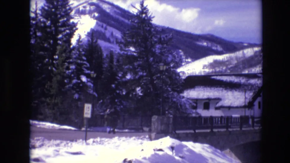 1974 Vail, Colorado 8mm frame showing the snowy roadside near the church, evergreen trees, mountain slopes, a bridge or railing, and a distant person walking.