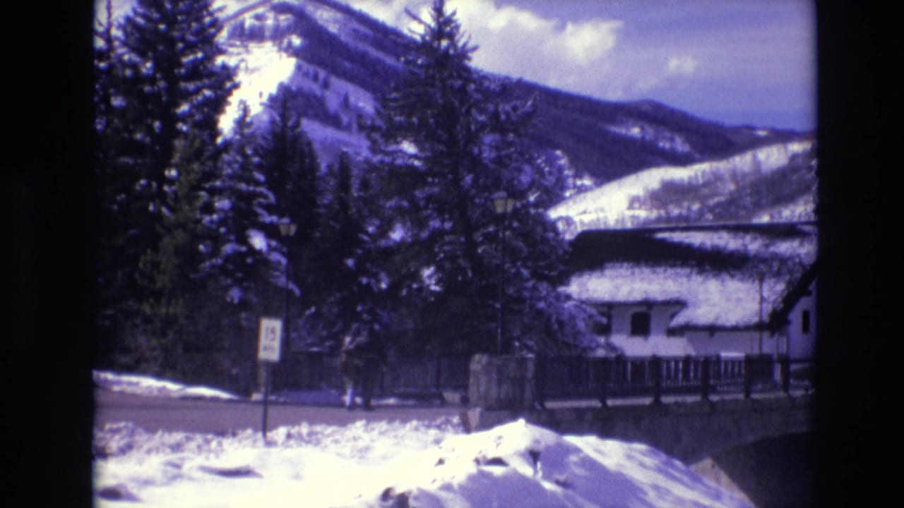 1974 Vail, Colorado home-movie frame showing a snow-covered roadside, evergreen trees, a white cabin or chalet, and mountain slopes in the background.