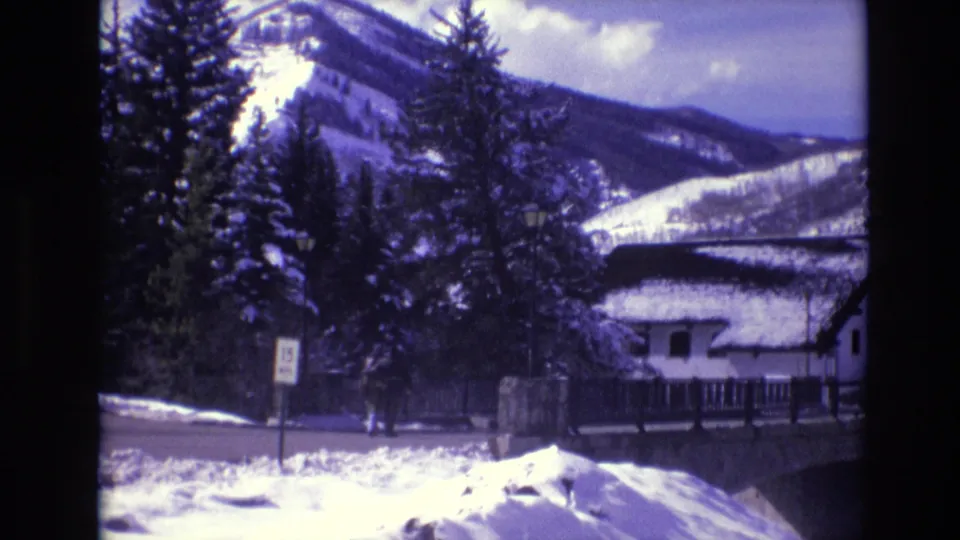 1974 Vail, Colorado home-movie frame showing a snow-covered roadside, evergreen trees, a white cabin or chalet, and mountain slopes in the background.