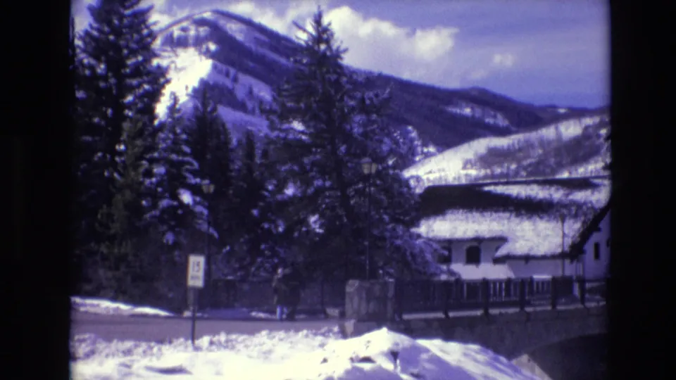 1974 Vail, Colorado frame with a snow-dusted cabin roof, dark evergreen trees, a roadside sign, and broad mountain slopes behind the village scene.