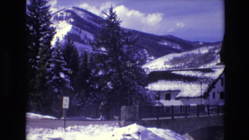 1974 Vail, Colorado home-movie frame showing a winter roadside with a small sign, evergreen trees, a white cabin, distant people, and snow-covered mountains.