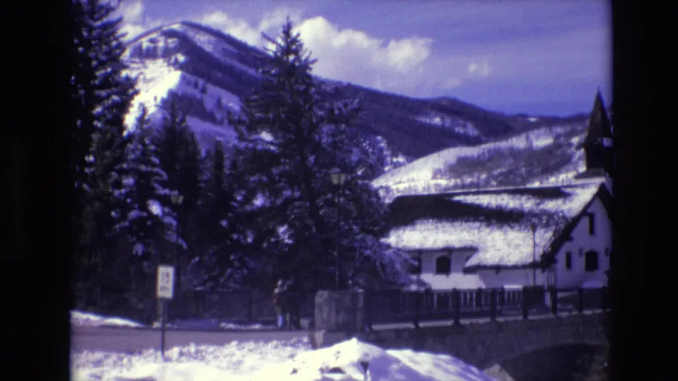 1974 Vail, Colorado frame with two distant people walking by a snow-covered roadside near a white cabin, evergreen trees, and mountain slopes.