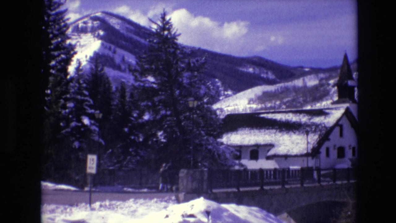 1974 Vail, Colorado 8mm frame showing snowy mountains behind evergreens, a white church-like building with a steeple, a bridge or wall, and a roadside sign.