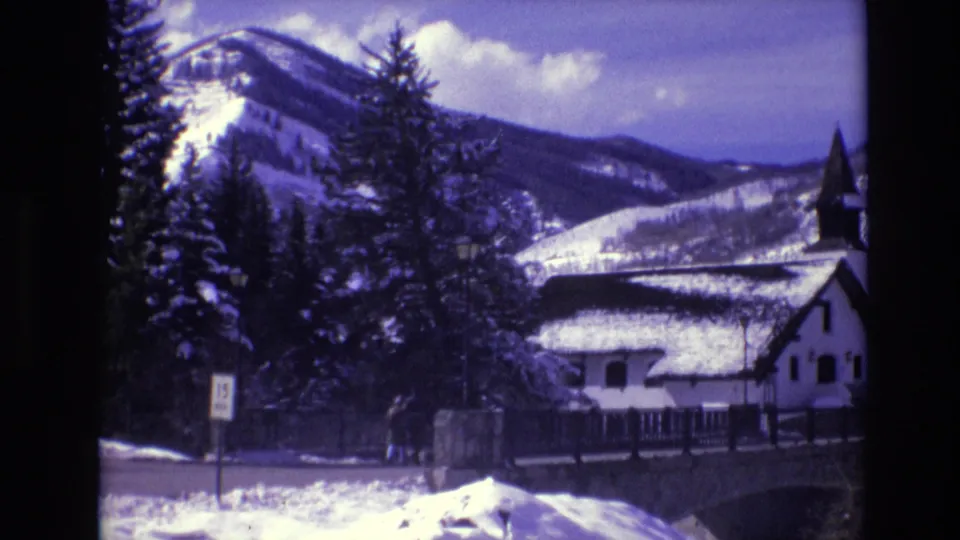1974 Vail, Colorado 8mm frame showing snowy mountains behind evergreens, a white church-like building with a steeple, a bridge or wall, and a roadside sign.