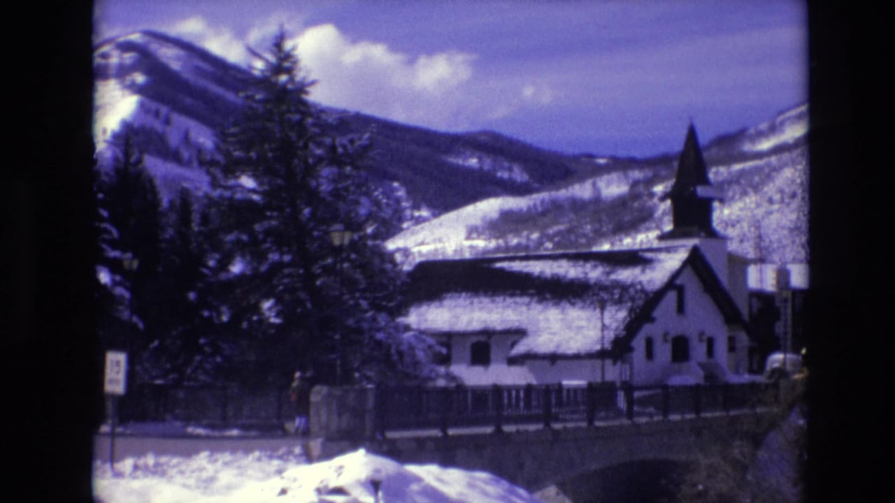 1974 Vail, Colorado home-movie frame of a snow-covered mountain backdrop, evergreen trees, a white steepled building, bridge structure, and roadside signage.