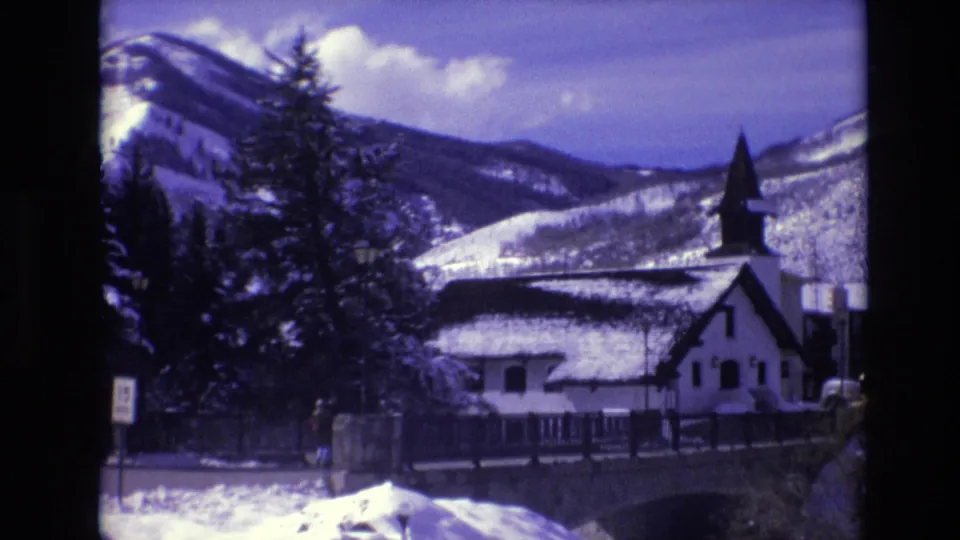 1974 Vail, Colorado home-movie frame of a snow-covered mountain backdrop, evergreen trees, a white steepled building, bridge structure, and roadside signage.